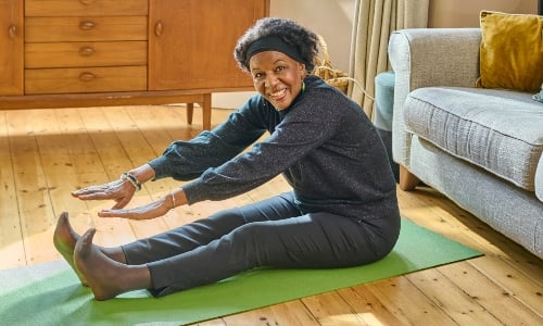 An older woman wearing workout gear, stretching on a yoga mat and smiling at the camera