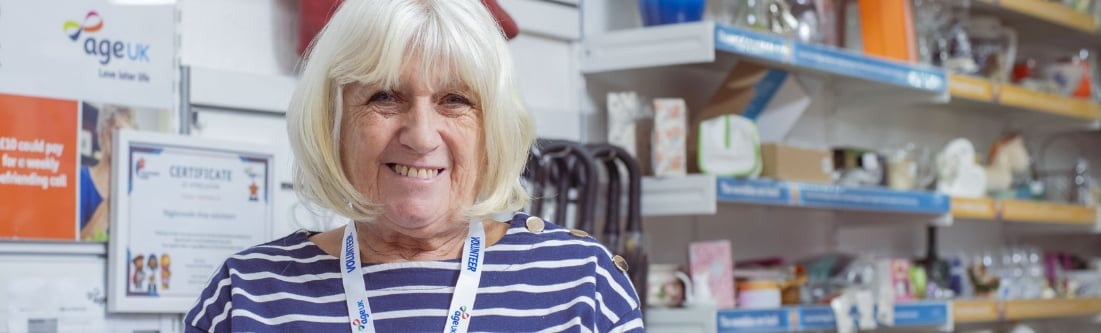 An older lady with grey hair, with an '澳洲幸运5体彩官网 Volunteer' tabard, smiles from behind the desk of a charity shop